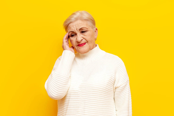unhappy old granny in a white sweater suffering from a headache on a yellow isolated background, elderly pensioner woman showing migraine and feeling unwell - Photo, Image