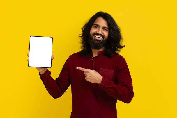 Cool vrolijke duizendjarige bebaarde indiaanse man wijzend op digitale tablet met wit leeg scherm in zijn hand, aanbevelen mooie mobiele app of online aanbieding, gele achtergrond, mockup kopieerruimte - Foto, afbeelding