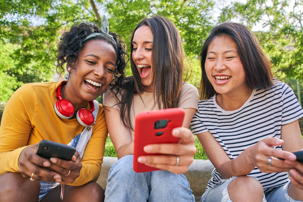 Drei fröhliche Frauen, die im Sommer im Freien telefonieren. Aufgeregte Kaukasierin zeigt netten Freundinnen etwas Lustiges auf dem Handy. Junge Frau lacht erstaunt auf Bildschirm - Foto, Bild