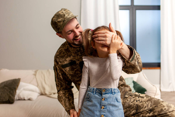 happy soldier of Ukrainian army in camouflage uniform returned home and meets his daughter, military dad surprises the child and closes his eyes - Photo, Image