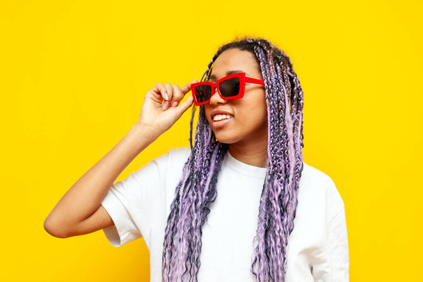 Hipster renkli rastalı ve sarı izole edilmiş arka planda güneş gözlüklü, eşsiz mor saç stili gözlüklü, gözlüklü genç Afro-Amerikalı kadın. - Fotoğraf, Görsel