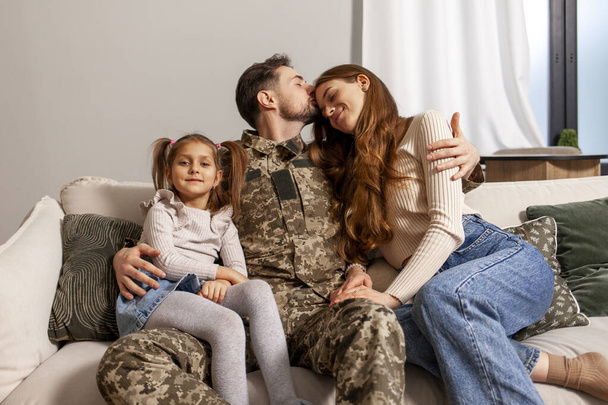 Oekraïense leger soldaat in camouflage uniform terug naar huis naar zijn familie, militaire cadet zit op de bank met zijn vrouw en dochter en glimlacht, kind knuffels veteranen vader - Foto, afbeelding