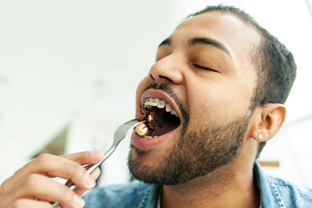 Hombre afroamericano con frenos comiendo pastel de chocolate con un tenedor, hombre sosteniendo tenedor con comida y mordiendo con la boca abierta, primer plano - Foto, imagen