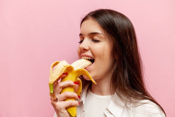 alegre mujer con frenos muerde plátano sobre rosa aislado fondo, chica joven come fruta, primer plano - Foto, imagen