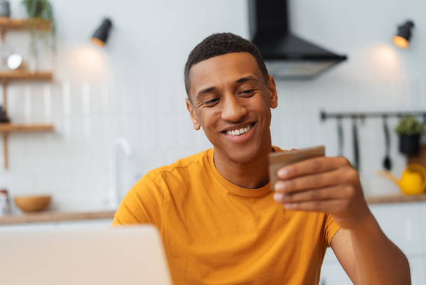 Boldog magabiztos afro-amerikai férfi kezében hitelkártya, laptoppal, választás, fizetés, online vásárlás. Jóképű férfi vásárol. Online banki koncepció - Fotó, kép