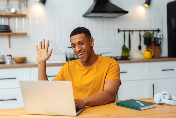 Portrait of smiling, happy young African American man sitting in modern kitchen at home, using laptop, talking on video call, waving hand. Online communication concept - Valokuva, kuva