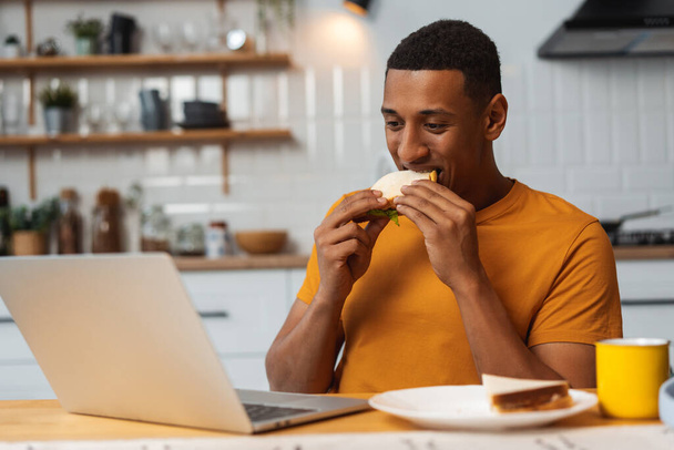 Uomo afroamericano attraente, positivo, libero professionista, lavoratore che tiene sandwich, mangia, utilizza il computer portatile mentre è seduto in cucina a casa. Bello studente che lavora online. Concetto di pausa, pranzo - Foto, immagini