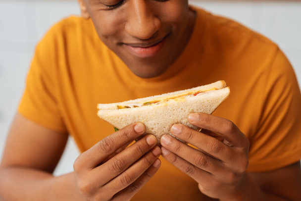 Πορτρέτο του όμορφου Αφροαμερικανού άνδρα κρατώντας σάντουιτς, απολαμβάνοντας το φαγητό, ενώ κάθεται στην κουζίνα στο σπίτι, closeup. Έννοια του γεύματος, των τροφίμων, δείπνο - Φωτογραφία, εικόνα