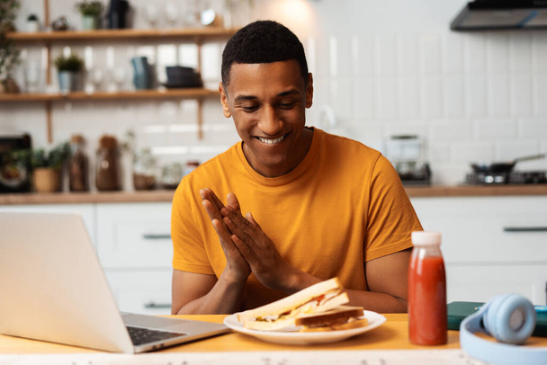 Retrato de un hombre afroamericano guapo sonriente que trabaja en casa sentado en la cocina, usando computadora portátil, merienda, sabroso sándwich. Trabajo remoto, concepto de comida - Foto, imagen