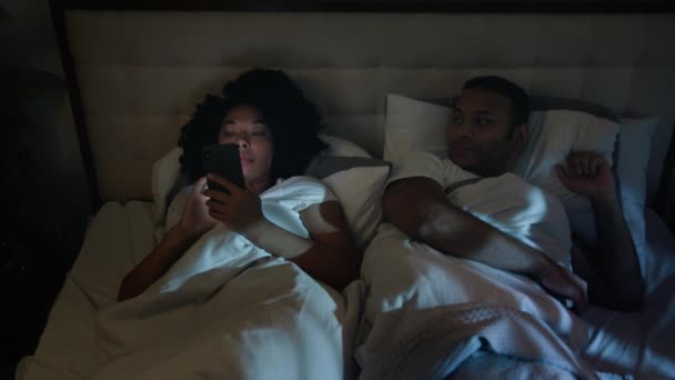 Afro-Amerikan cihaz bağımlısı sosyal medya cep telefonu bağımlısı kadın gece yatakta akıllı telefon kullanıyor üzgün, mutsuz adam, boşanmış koca, uyumaya çalışıyor. - Video, Çekim