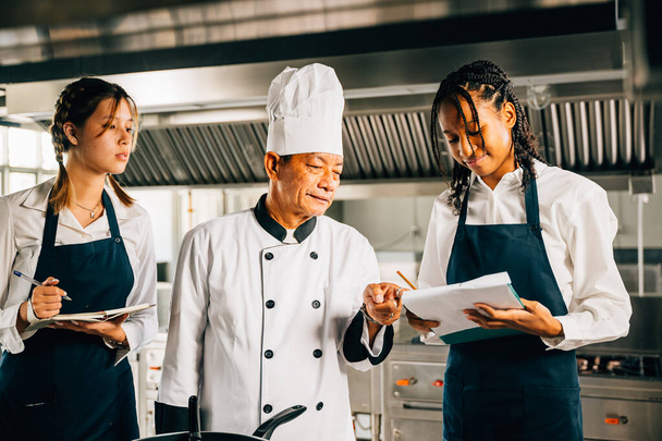 Professional Asian senior chef guides diverse students in a restaurant kitchen workshop. Focusing on teamwork learning and note-taking. Educational environment. Food Edocation - Fotoğraf, Görsel