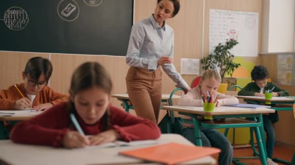 Caucasian woman teacher in elementary school teach children class kids writing task in classroom knowledge studying primary education lesson female tutor teaching learning boys girl study pupils learn - Footage, Video