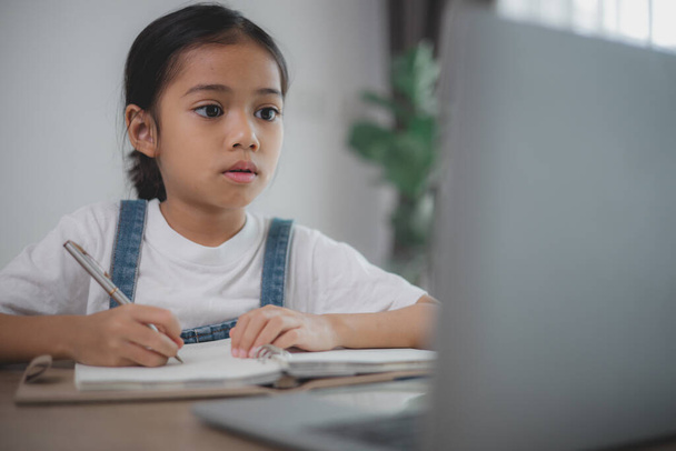 Ein junges Mädchen sitzt an einem Schreibtisch mit Laptop und Notizbuch. Sie schreibt in das Notizbuch und blickt auf den Laptop. Die Szene legt nahe, dass sie an einem Projekt arbeitet oder für eine Prüfung studiert. - Foto, Bild