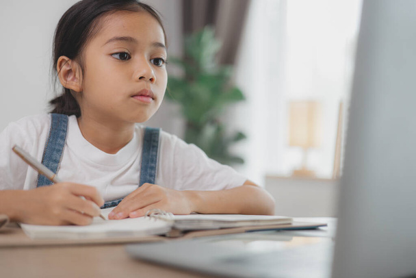 Uma jovem está sentada em uma mesa com um laptop aberto na frente dela. Ela está escrevendo em um caderno - Foto, Imagem