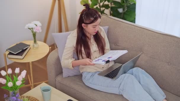 Azjatycka bizneswoman pracująca w domu, korzystająca z laptopa na kanapie w salonie. - Tak. Wysokiej jakości materiał 4k - Materiał filmowy, wideo