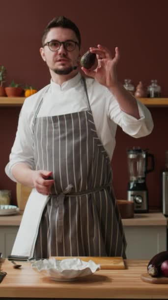 Κάθετη μεσαία μαχαιριά του Καυκάσου σεφ σε ενδύματα εργασίας και ποτήρια στέκεται από ξύλινο τραπέζι μαγειρικής στην κουζίνα και δείχνει πώς να κόψει ώριμο πράσινο αβοκάντο στην κάμερα - Πλάνα, βίντεο
