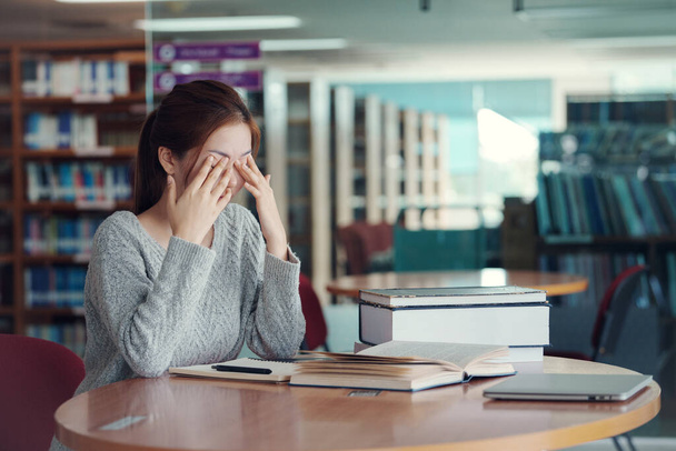 Junge asiatische Studentin reibt sich die Augen, fühlt sich müde, nachdem sie ein Buch in der Bibliothek gelesen hat. - Foto, Bild