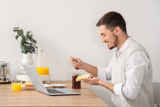 Laptoplu genç adam mutfakta lezzetli reçelli tost yiyor. - Fotoğraf, Görsel