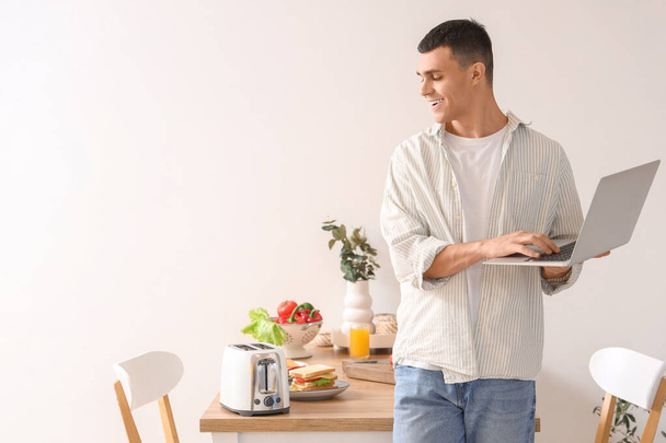 Young man using laptop while waiting for toasts in toaster in kitchen - Photo, Image