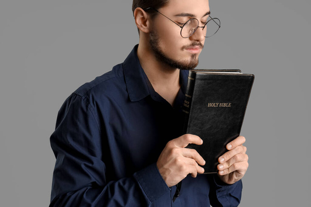 Молодий чоловік з Біблією молиться на сірому фоні - Фото, зображення