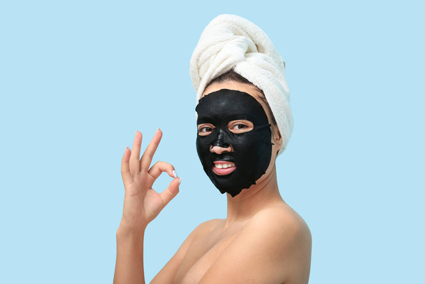 Νεαρή γυναίκα με μάσκα ενεργού άνθρακα στο πρόσωπό της δείχνει ΟΚ σε μπλε φόντο - Φωτογραφία, εικόνα