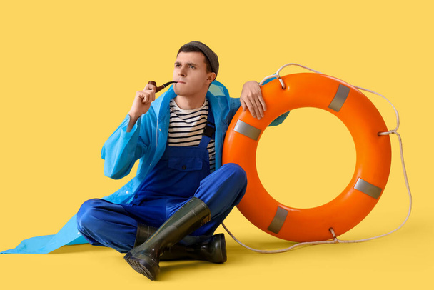 Молодой моряк в плаще с курительной трубкой и кольцевым буем на желтом фоне - Фото, изображение