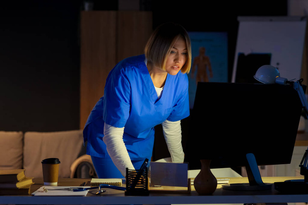 Женщина-медсестра с компьютером работает в вечернюю смену за столом в клинике - Фото, изображение