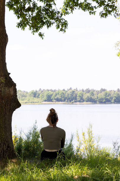 Egy derűs kép egy nőről, aki egy lombos fa alatt ül, és egy nyugodt tó felett néz ki. A természetes lombkorona és a fa szelíd ölelése békés elmélkedés légkörét teremti meg. - Fotó, kép