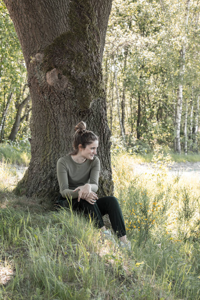 Dit beeld vangt een jonge vrouw die geniet van een rustig moment in het bos, zittend aan de voet van een grote boom met zonlicht dat door de bladeren stroomt, wat een vredige en reflecterende sfeer creëert. - Foto, afbeelding