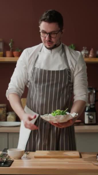 Vertikální střední snímek mladého kavkazského šéfkuchaře s mikrofonem popisujícím a zobrazujícím proces přípravy salátu před kamerou, sypání semen na něm při dokončování - Záběry, video