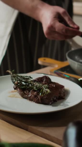 Vertical cropped shot of unrecognizable male chef serving two delicious beef steaks with thyme and garlic on white plate - Footage, Video