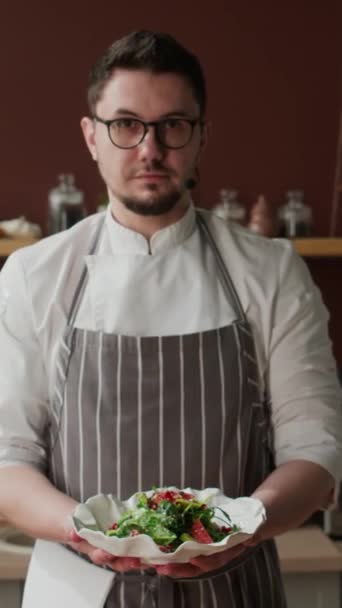 Portrait vertical moyen du chef caucasien portant des lunettes, un manteau de chef et un tablier rayé posant pour appareil photo avec une délicieuse salade verte dans une assiette en céramique blanche dans les mains dans la cuisine - Séquence, vidéo