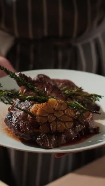 Vertikální detailní záběr nerozpoznatelného pánského šéfkuchaře pózujícího se dvěma šťavnatými pánvičkami smaženého hovězího steaku na bílém talíři - Záběry, video