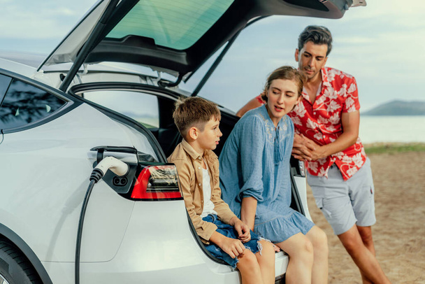 Voyage de vacances en famille voyageant par la plage avec voiture électrique, belle famille assis sur le coffre, charge batterie de voiture EV avec l'énergie verte et durable. Voyage en famille et voiture écologique. Perpétuel - Photo, image