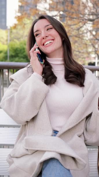 Κάθετα. Μια μελαχρινή κυρία κάνει μια κλήση με ένα κινητό τηλέφωνο κάθεται σε ένα παγκάκι σε ένα δρόμο της πόλης μετά τα ψώνια. Απομονωμένη νεαρή γυναίκα. Χαμογελώντας και μιλώντας με ένα smar tphone. Επικοινωνία. Αργή κίνηση - Πλάνα, βίντεο