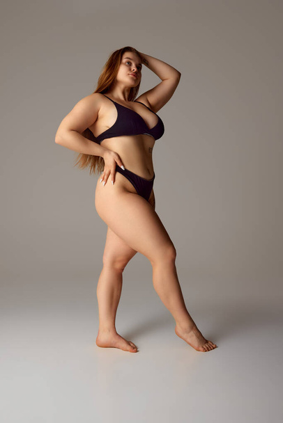 Mujer joven atractiva y confiada posando en lencería negra, bikini mirando hacia otro lado contra fondo gris del estudio. Concepto de belleza natural, feminidad, positividad corporal, dieta, fitness. - Foto, imagen