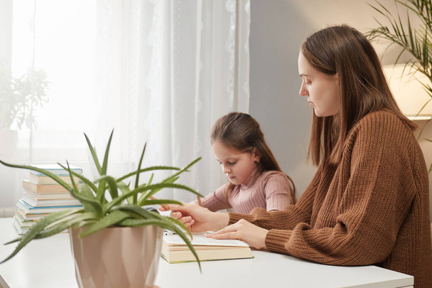 Brown haired mother and daughter learning to write woman teaches child helps with homework while sitting at table in living room kids learning at home - Foto, Imagem