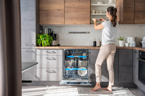 Жінка видаляє чистий керамічний посуд з посудомийної машини. Концепція побутової та корисної технології. Високоякісна фотографія.  - Фото, зображення