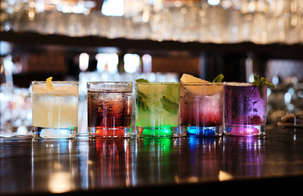 Een verscheidenheid van vijf kleurrijke cocktails, elk uniek versierd, zijn opgesteld op een donker houten bar teller, verlicht van onderen. De glazen reflecteren omgevingslicht, creëren een elegante en uitnodigende sfeer. - Foto, afbeelding