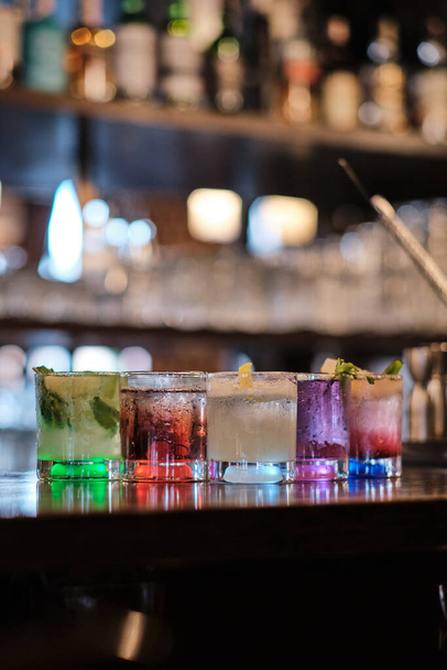 Erilaisia viisi värikästä cocktaileja, kukin ainutlaatuisesti koristeltu, on rivissä tumma puinen baari laskuri, valaistu alhaalta. Lasit heijastavat ympäristön valoa, mikä luo tyylikkään ja kutsuvan ilmapiirin.. - Valokuva, kuva