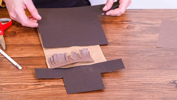 Schritt für Schritt. Der Lehrer führt den Online-Unterricht durch die Herstellung einer Papierpuppe aus einer braunen Tasche, wobei er eine Holzoberfläche kreativ als Arbeitsplatz nutzt. - Foto, Bild
