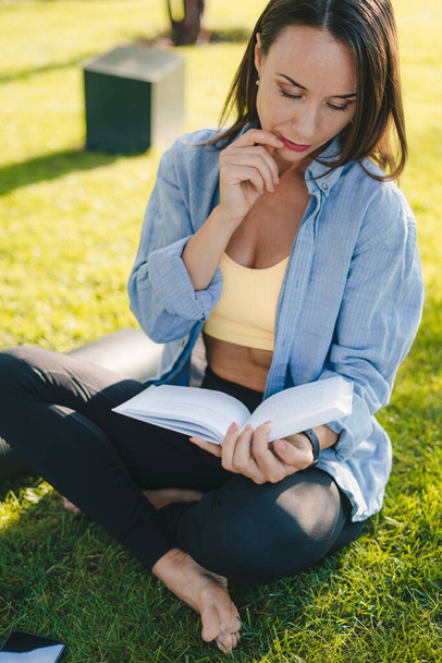Attractive adult woman reading interesting book sitting on green grass at city park in sunny day. Enjoying free time concept. - Photo, Image