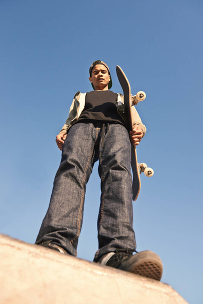 Скейтпарк, серйозний і портрет людини з скейтбордом, що практикує змагання з навичками. Фітнес, хобі та вид знизу молодого ковзаняра на рампі для тренувань на відкритому повітрі з блакитним небом - Фото, зображення