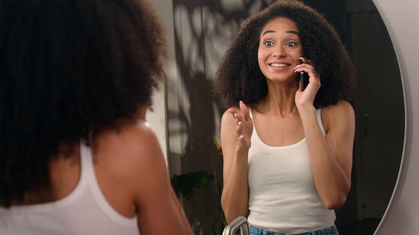 Happy African woman girl talking mobile phone laughing talk speaking smartphone call friend pleasant conversation looking at bathroom mirror check hair checking fix hairdo hairstyle laugh gossiping - Photo, Image