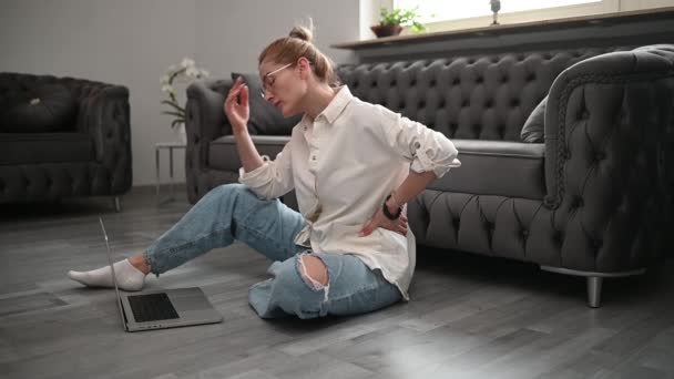 Eine blonde Frau mit Brille sitzt auf dem Boden neben dem Sofa und arbeitet zu Hause mit Laptop. Hochwertiges FullHD-Filmmaterial - Filmmaterial, Video