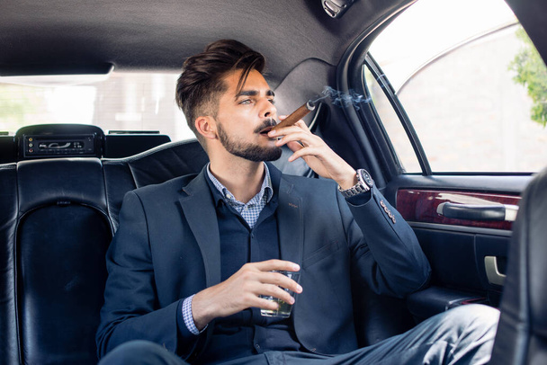 Young business man enjoying cigar and drink in limousine - Photo, Image