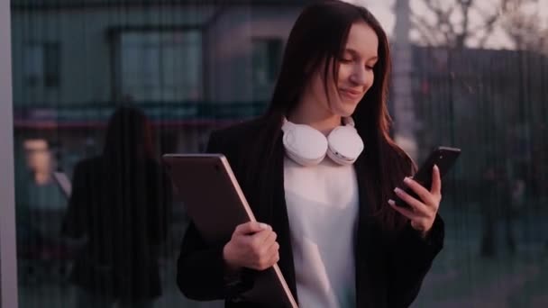 A businesswoman holds a laptop in one hand and a phone in the other, resolving work matters over the phone. Office worker, freelancer, social media manager, outdoor work. - Footage, Video
