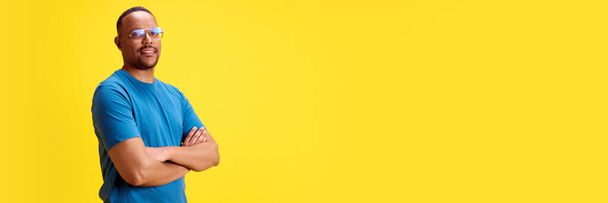 Confident, smiling African-American man standing with hands crossed against yellow studio background. Success. Concept of human emotions, casual fashion, lifestyle. Banner. Empty space for text, ad - Fotó, kép