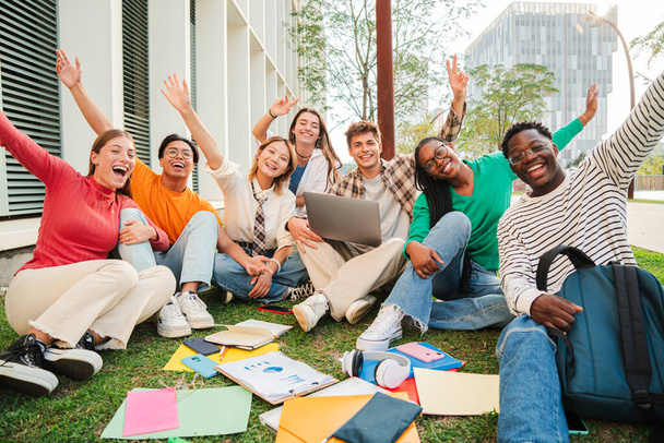 Portret dużej grupy szczęśliwych nastolatków siedzących na trawniku kampusu uniwersyteckiego, gestykulujących i celebrujących wspólnie cel akademicki. Pozytywne wielorasowe nastolatki gestykulujące i patrzące w kamerę - Zdjęcie, obraz