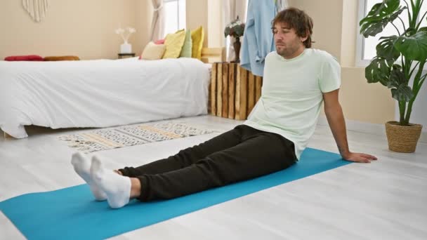 Een ontspannen jongeman zit op een yogamat in een lichte home bedroom setting, omgeven door gezellige inrichting. - Video